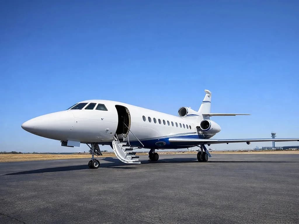 A white and blue Dassault Falcon business jet parked on a tarmac under a clear blue sky with its door open and stairs extended.
