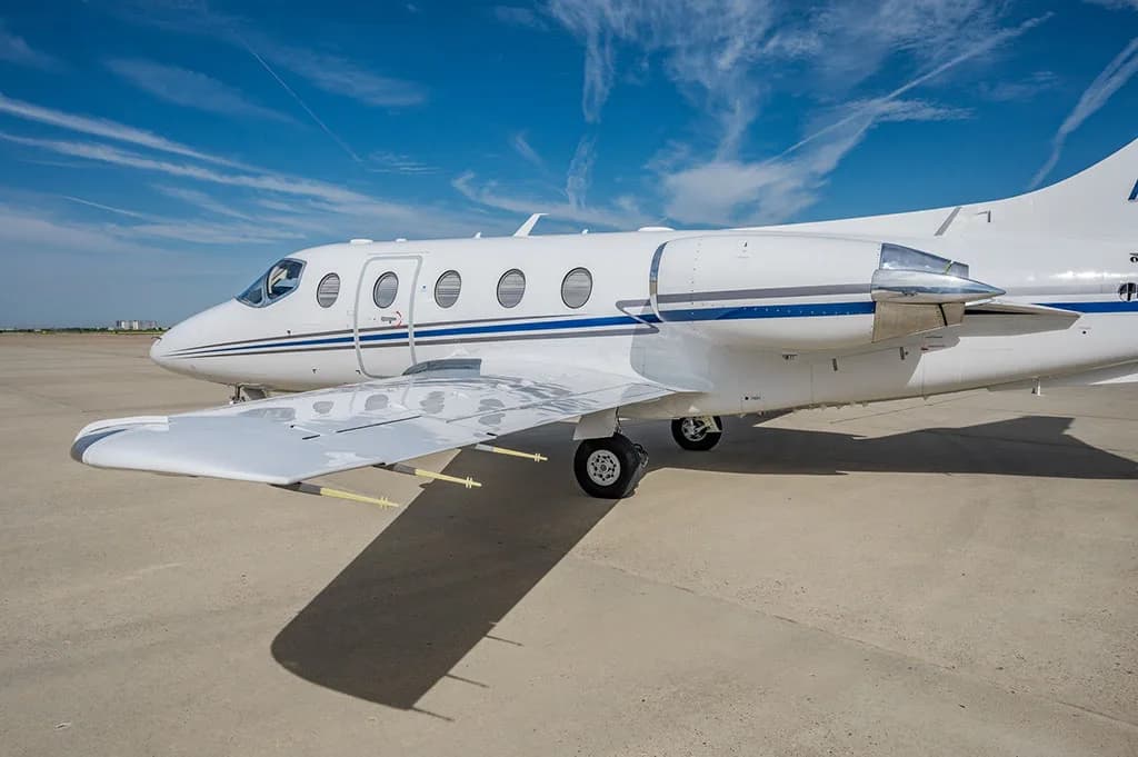 A close-up view of the mid-fuselage, wing, and engine of a white business jet with blue stripes on a tarmac.