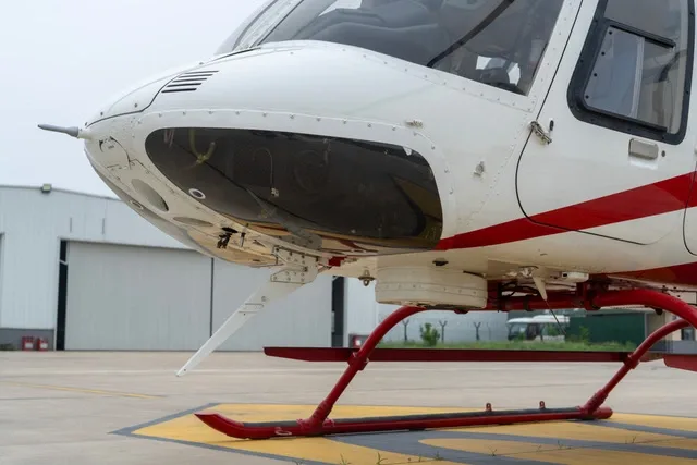 A close-up view of the nose and red landing skids of a white and red helicopter on an airfield with a hangar in the background.