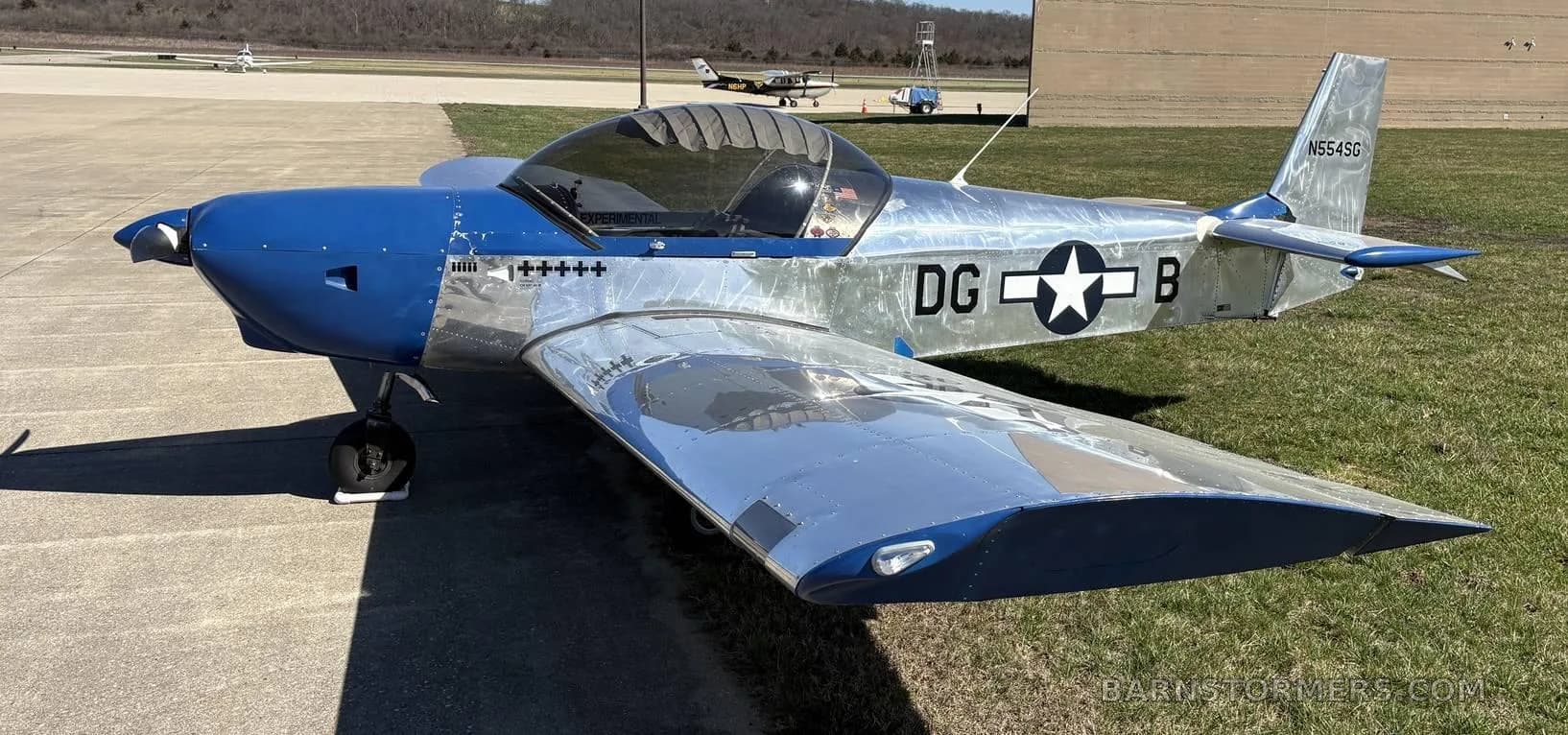 A blue and polished aluminum single-engine aircraft with a bubble canopy and "DG * B" markings, viewed from the front-right on an airfield.