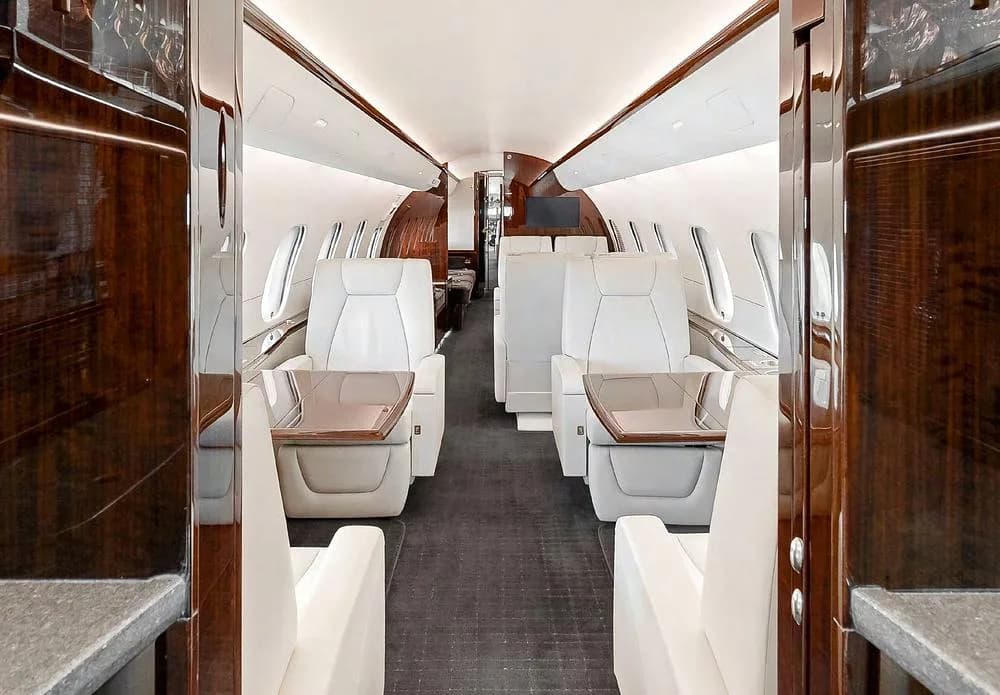 View down the aisle of a private jet cabin featuring cream seats, dark wood paneling, and a dark carpet.