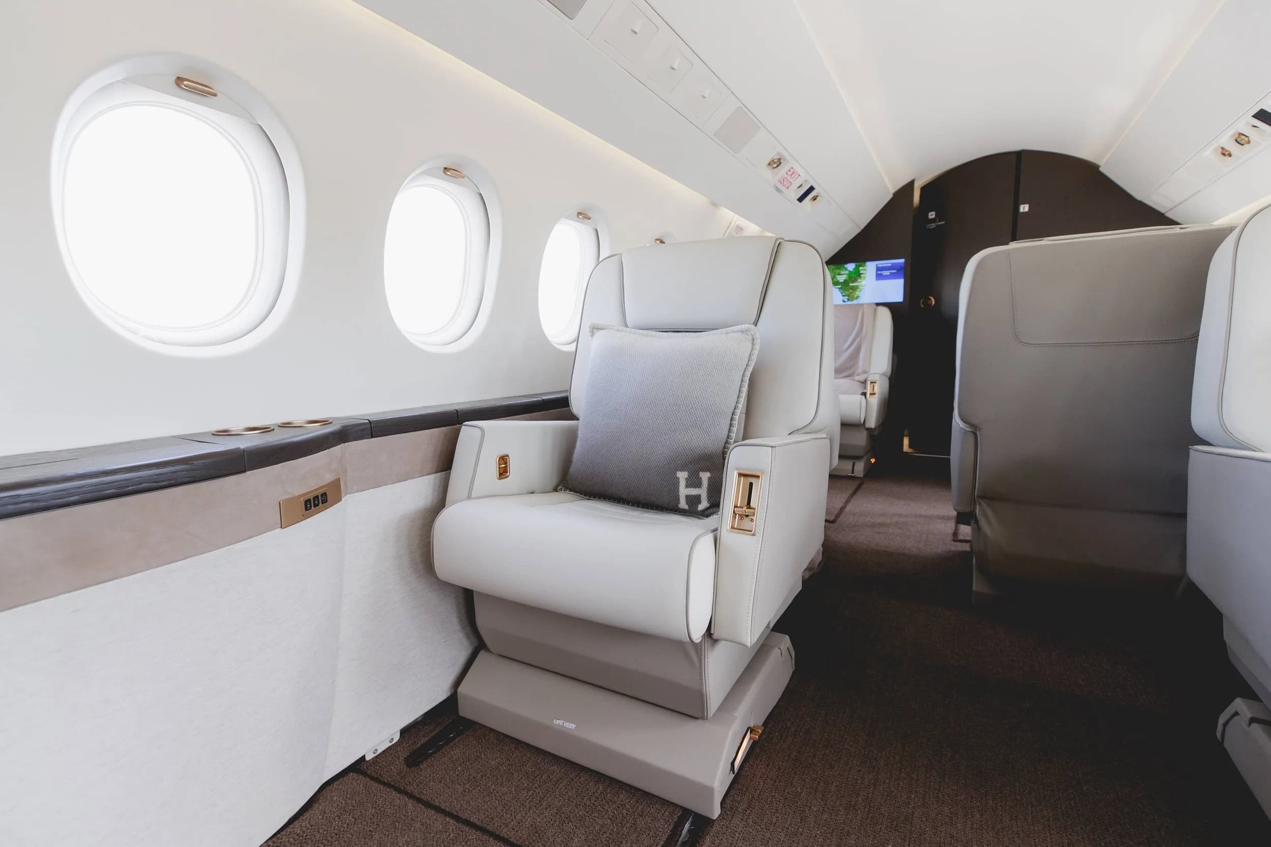 A close-up view of a white leather aircraft seat with an "H" monogrammed pillow and a window.