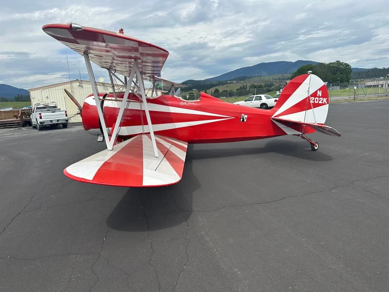 A red and white biplane with N202K on the tail, parked on an asphalt apron with mountains in the background.