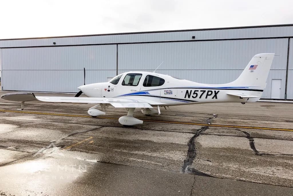 A side view of a white Cirrus SR22 aircraft with blue and black stripes, tail number N57PX, parked on a wet airport tarmac.