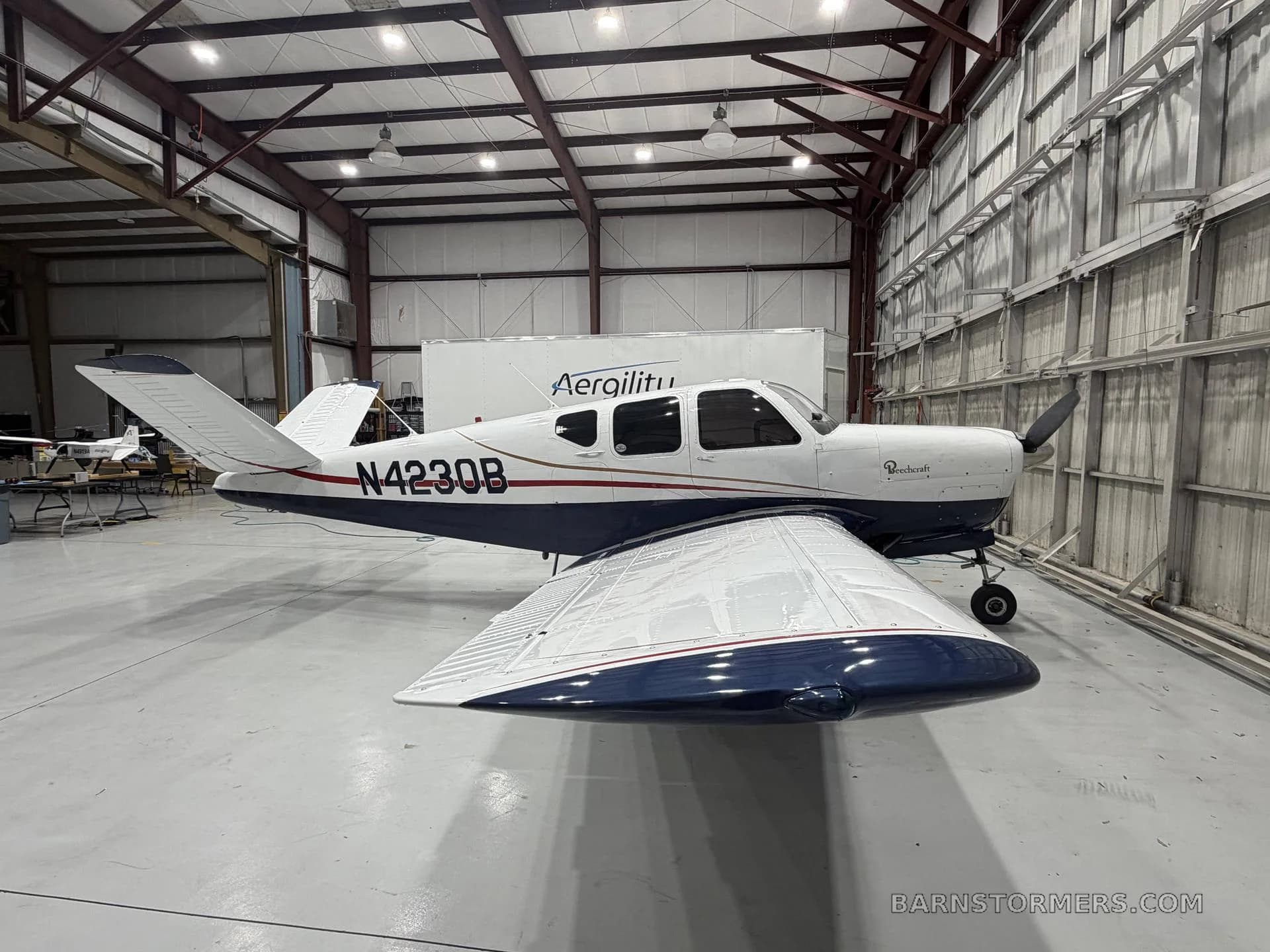 A white and blue Beechcraft Bonanza (N4230B) is parked inside a hangar, viewed from the front-left.