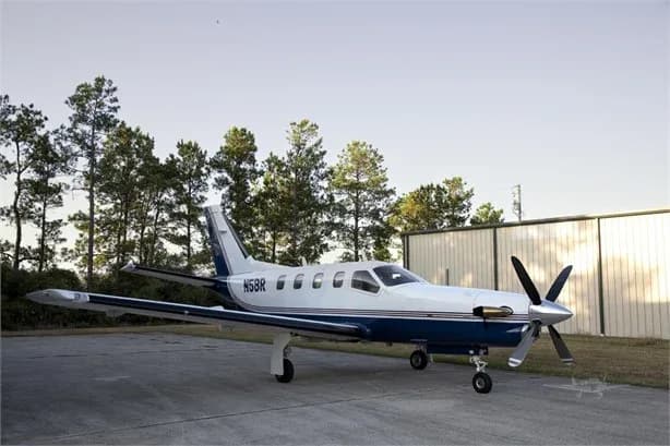 A white and blue single-engine turboprop aircraft, N5BR, is parked on an asphalt ramp with trees and hangars in the background.
