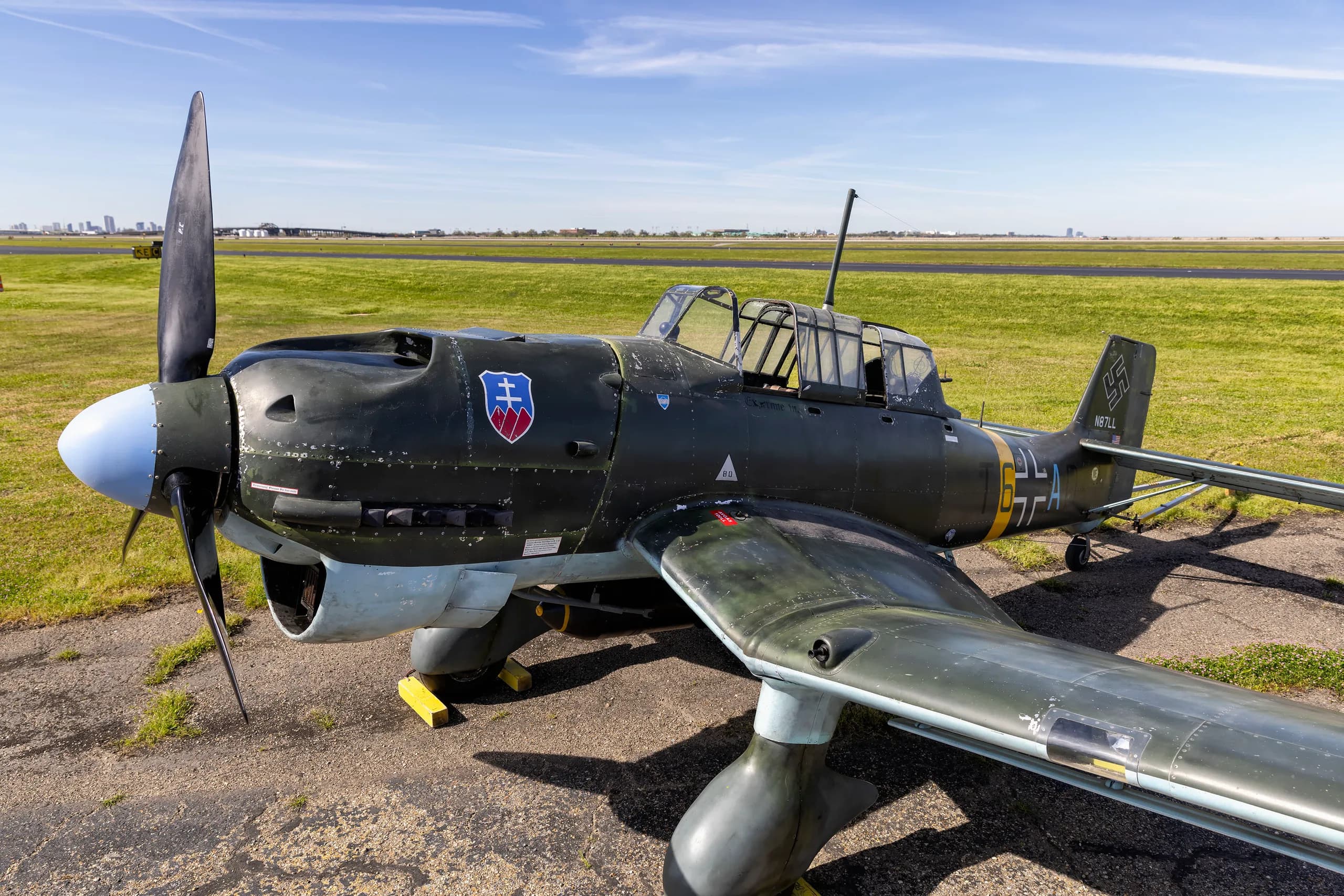 A side view of a dark green Junkers Ju 87 Stuka with an open canopy, parked on an airfield.