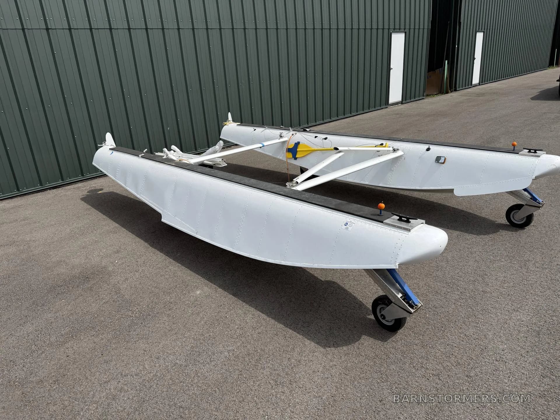 A side view of two white aircraft floats with wheels parked on an asphalt apron next to a green hangar.