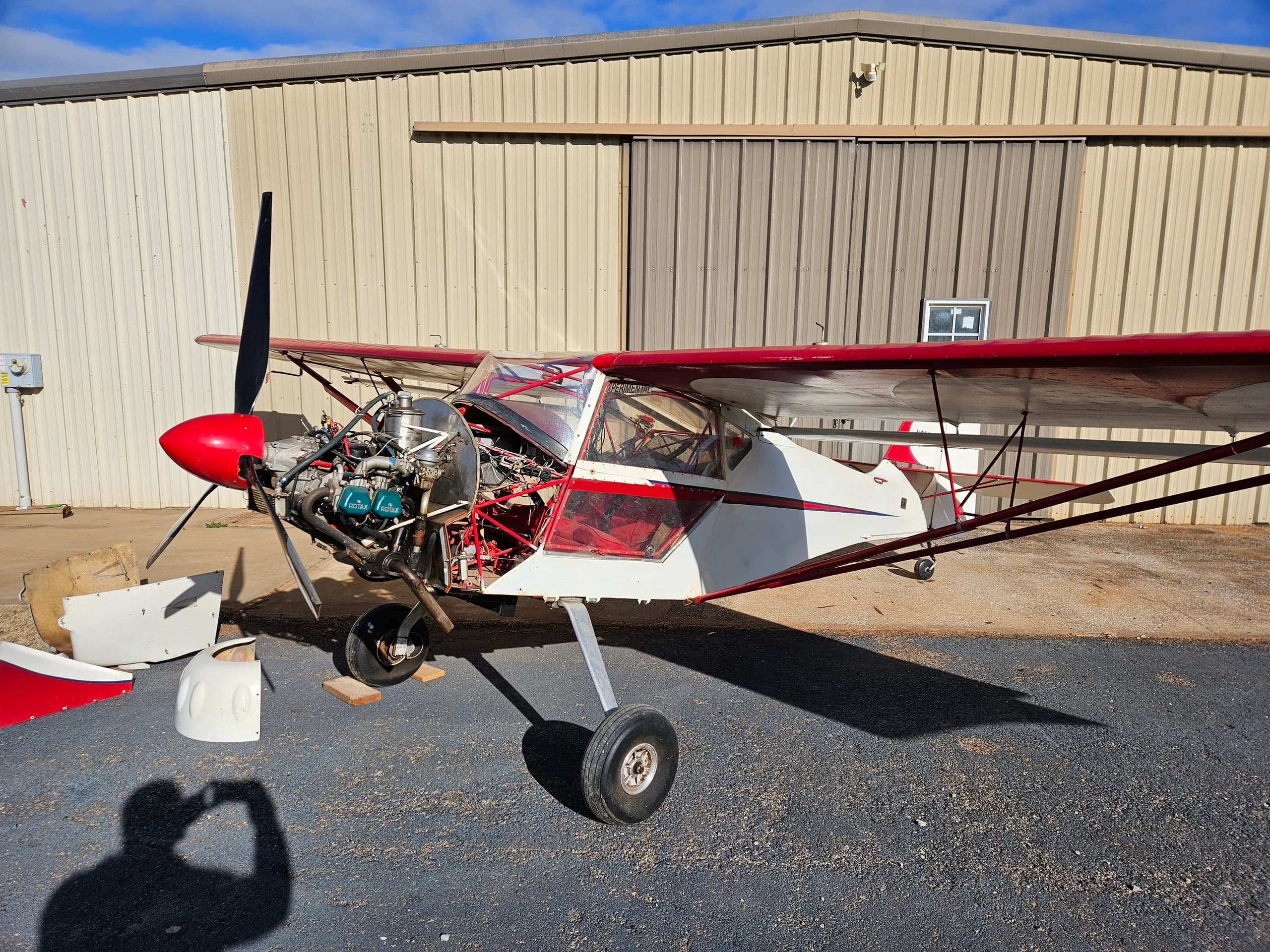 A close-up view of the front left side of a white and red experimental aircraft with its engine cowling removed, showing the exposed engine and propeller.