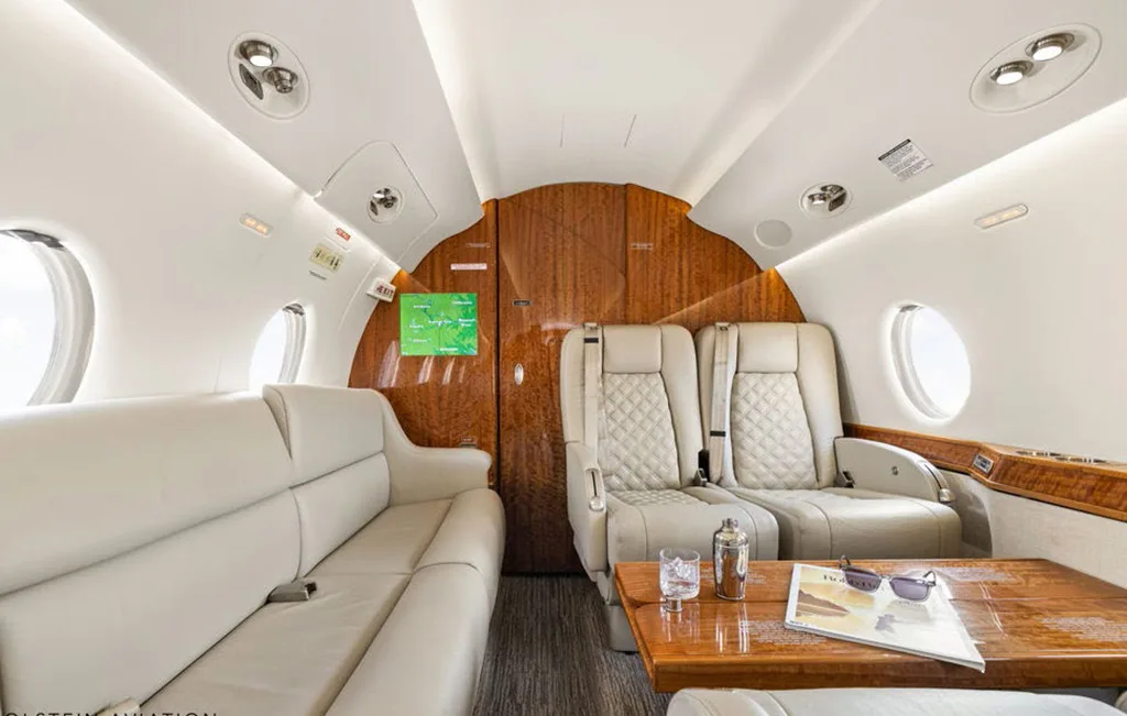 The spacious interior of a private jet cabin features a long beige sofa and two individual seats with a wooden table.
