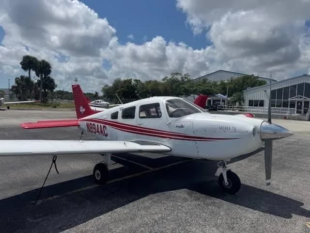 A white Piper Archer TX with red stripes and tail, registration N894AC, parked on an airport tarmac with hangars in the background.