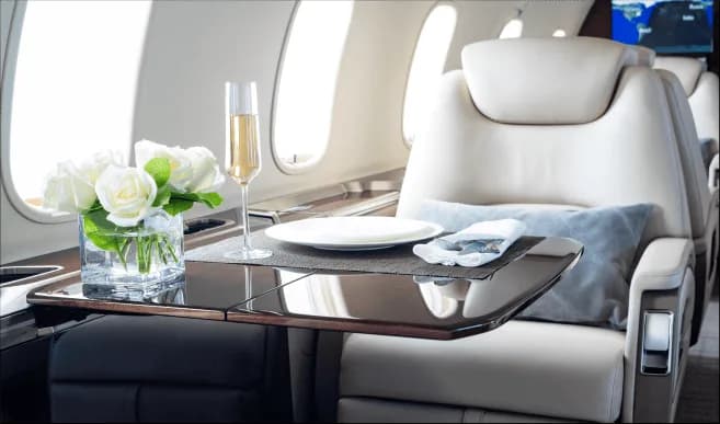 A private jet cabin table is set with a plate, silverware, a glass of champagne, and a vase of white roses.