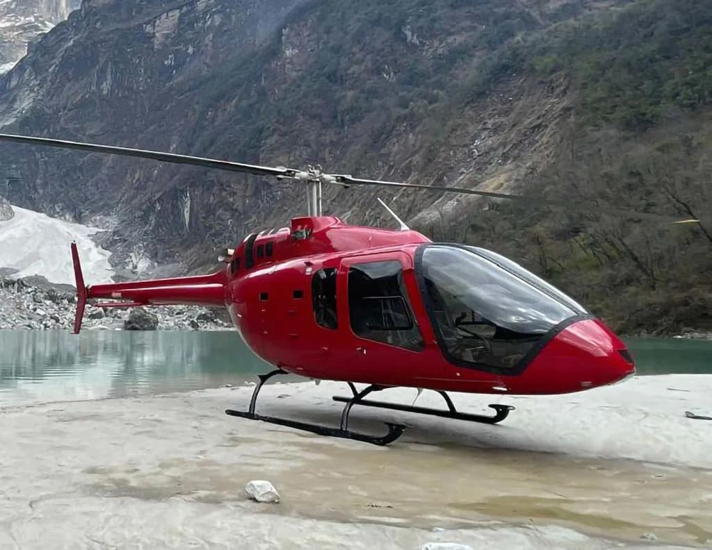 A red helicopter with black skids sits on a rocky shore next to a turquoise lake, with mountains in the background.