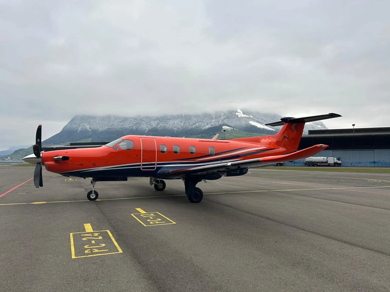 A bright orange Pilatus PC-12 aircraft with dark blue and white stripes sits on an airport tarmac with snow-capped mountains in the background.