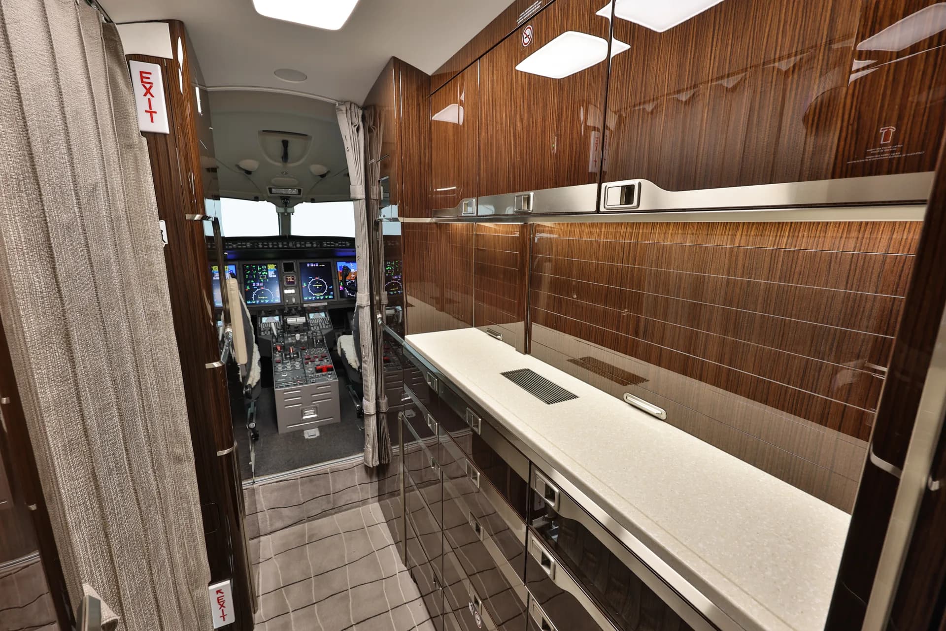 A view from the aircraft galley with dark wood finishes, looking forward into the cockpit with its illuminated displays.