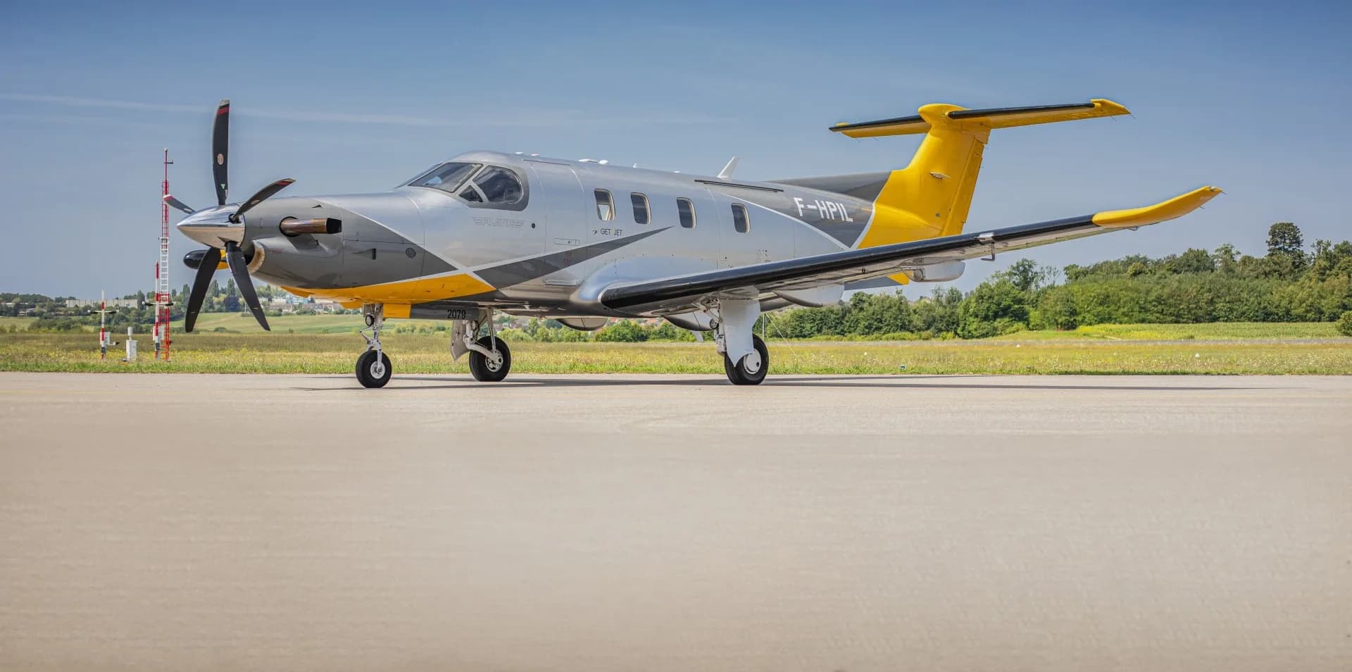 A silver and yellow Pilatus PC-12 aircraft (F-HPIL) parked on an airfield tarmac under a clear blue sky.