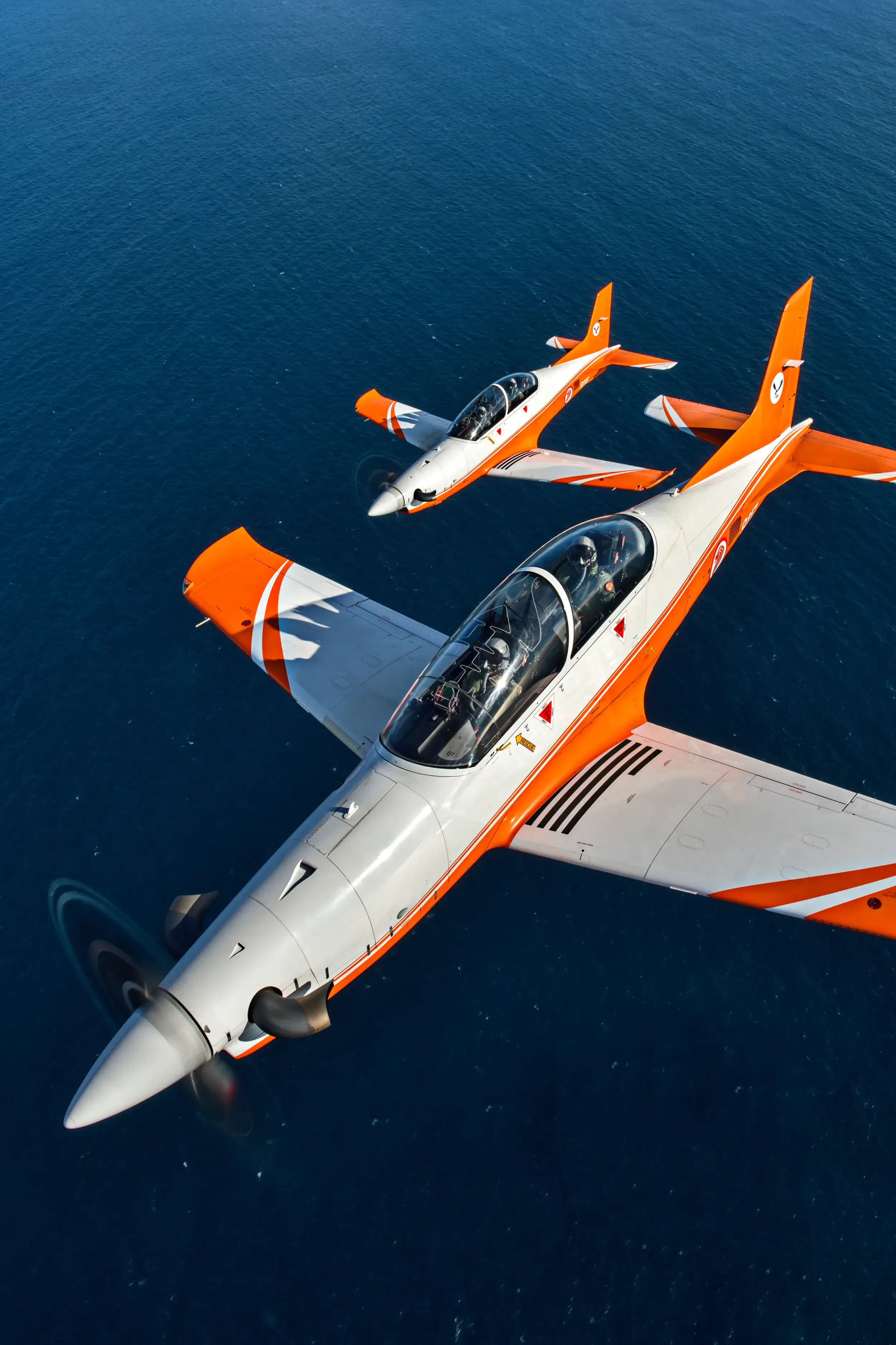 Two orange and white Pilatus PC-21 aircraft flying in formation over the ocean.