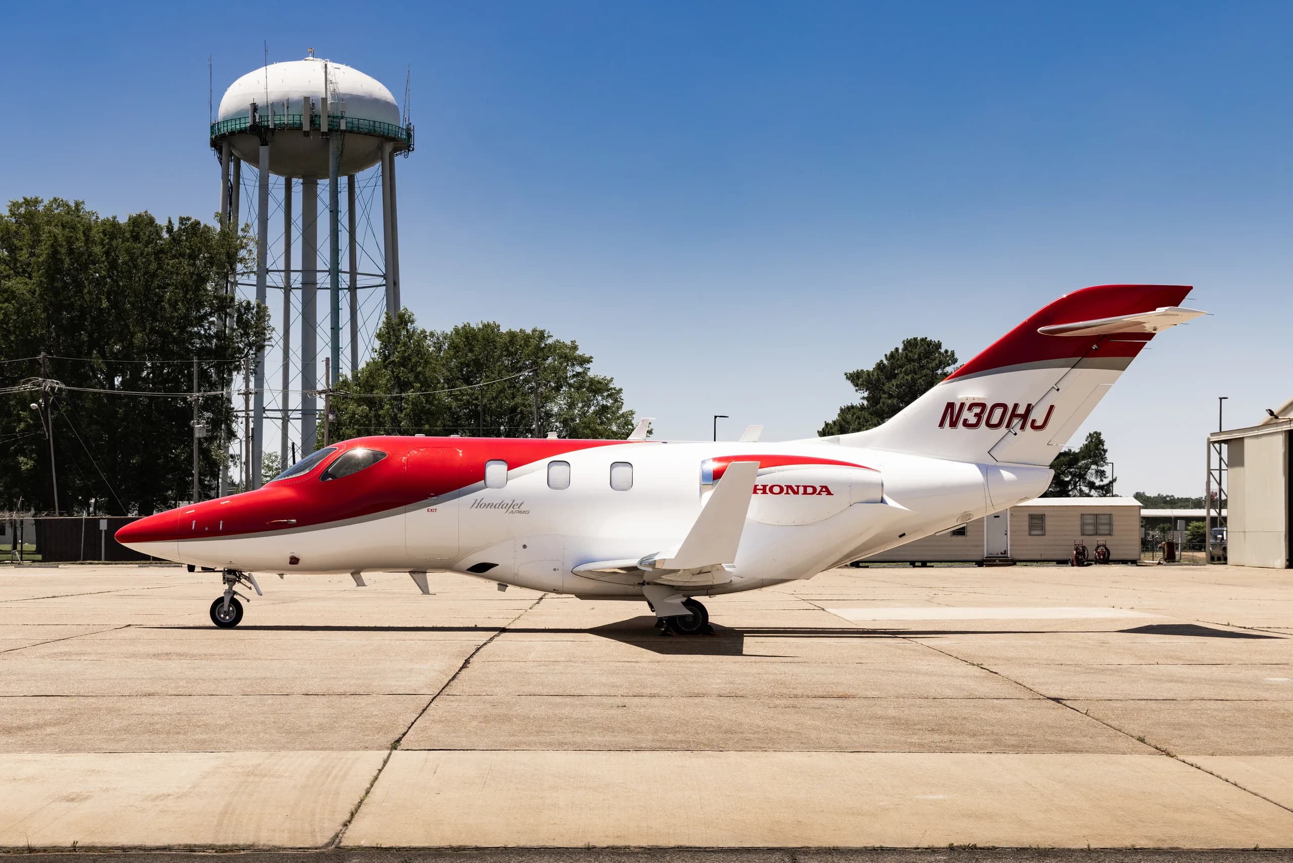 A white and red HondaJet HA-420 (N30HJ) parked on a tarmac with a water tower in the background, viewed from the left side.