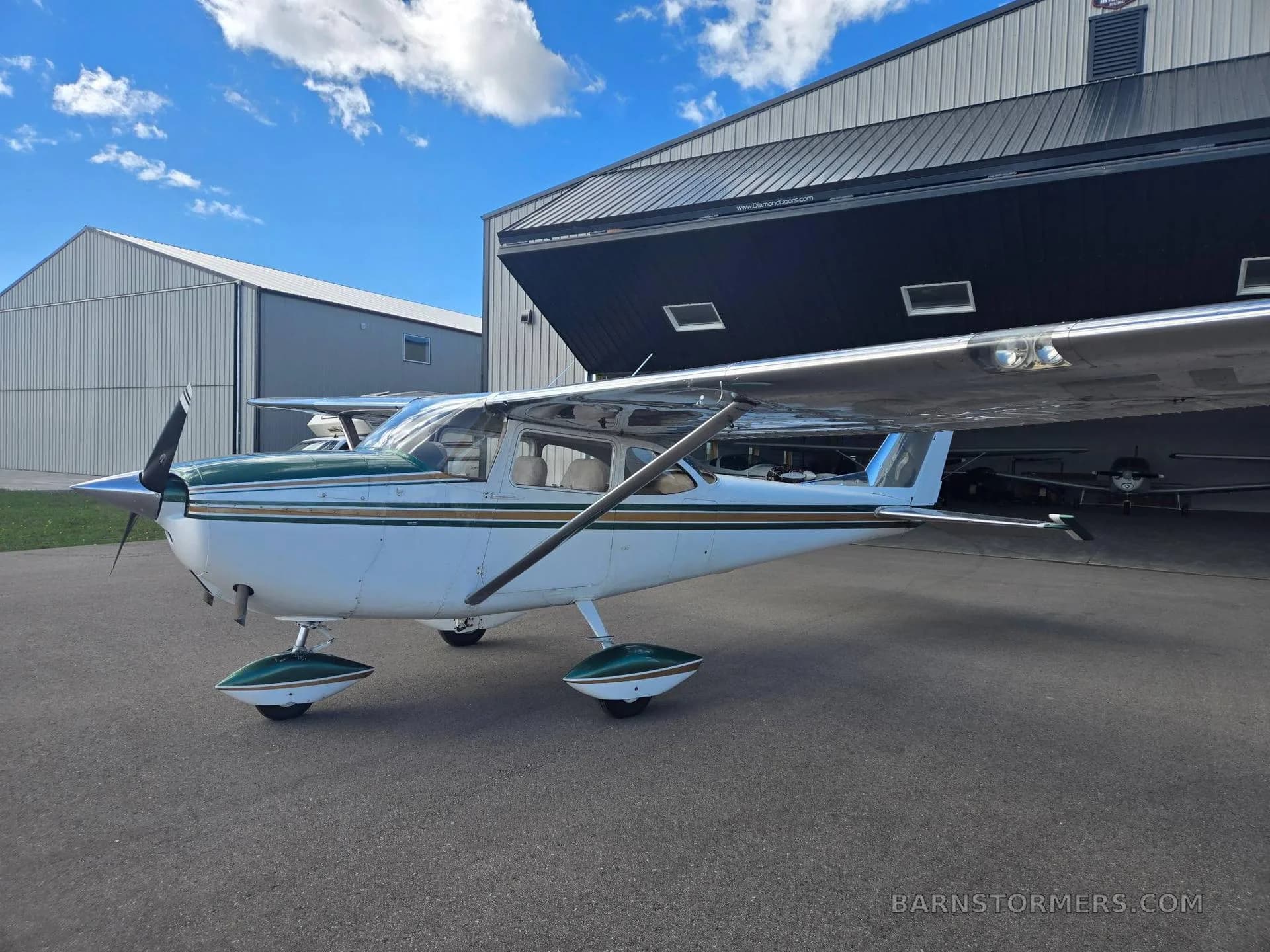 A white and green Cessna 172 is parked on the tarmac next to an open hangar under a blue sky.