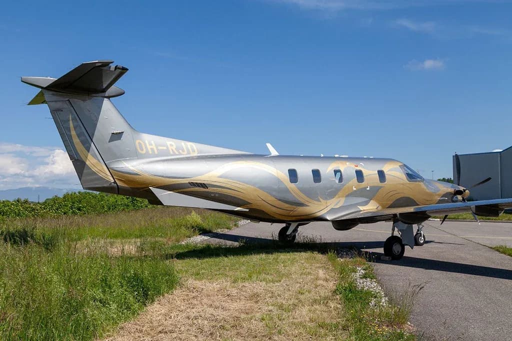 A rear-side view of a silver and gold Pilatus PC-12 aircraft (OH-RJD) parked on an airfield next to grass.