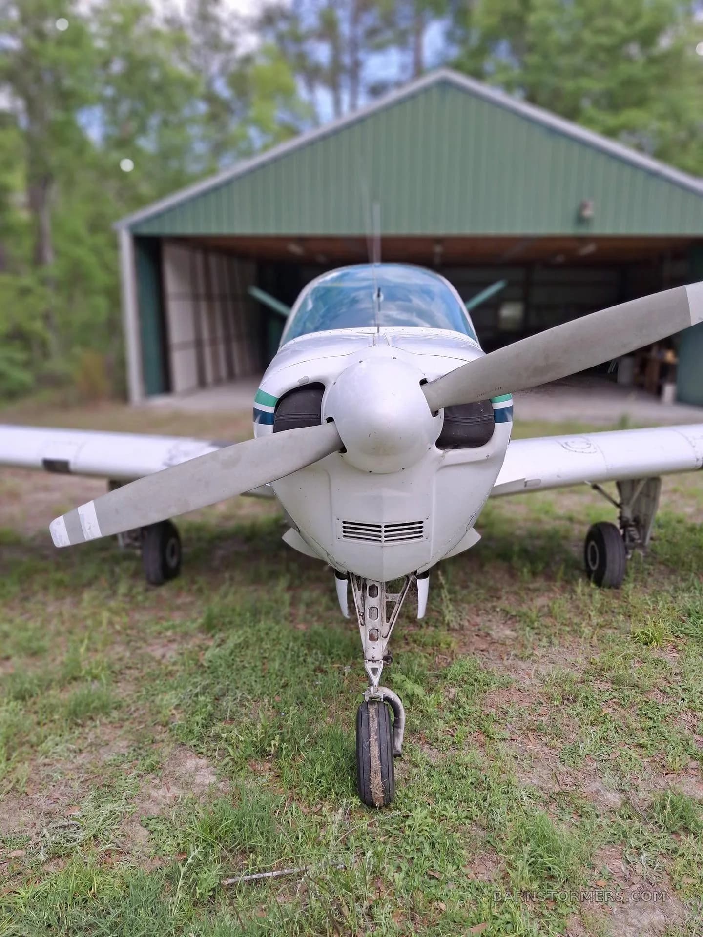 A front view of a white single-engine propeller aircraft with green stripes, parked on grass in front of a green hangar.
