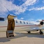 A white private jet with a red stripe is parked on an tarmac with its main door open, under a blue sky with clouds.