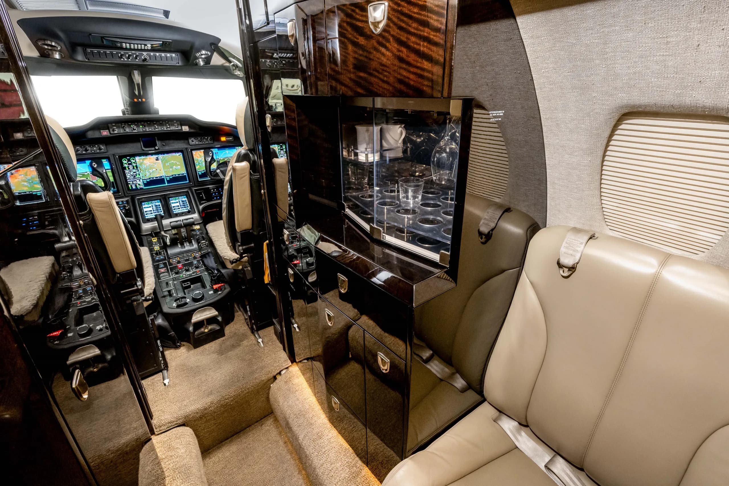 The galley area of a business jet with a dark wood cabinet and glassware, looking towards the cockpit.