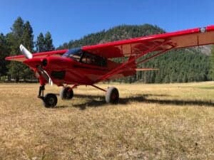 A red Cubcrafters XCub aircraft parked on a grassy airstrip with trees and a mountain in the background.