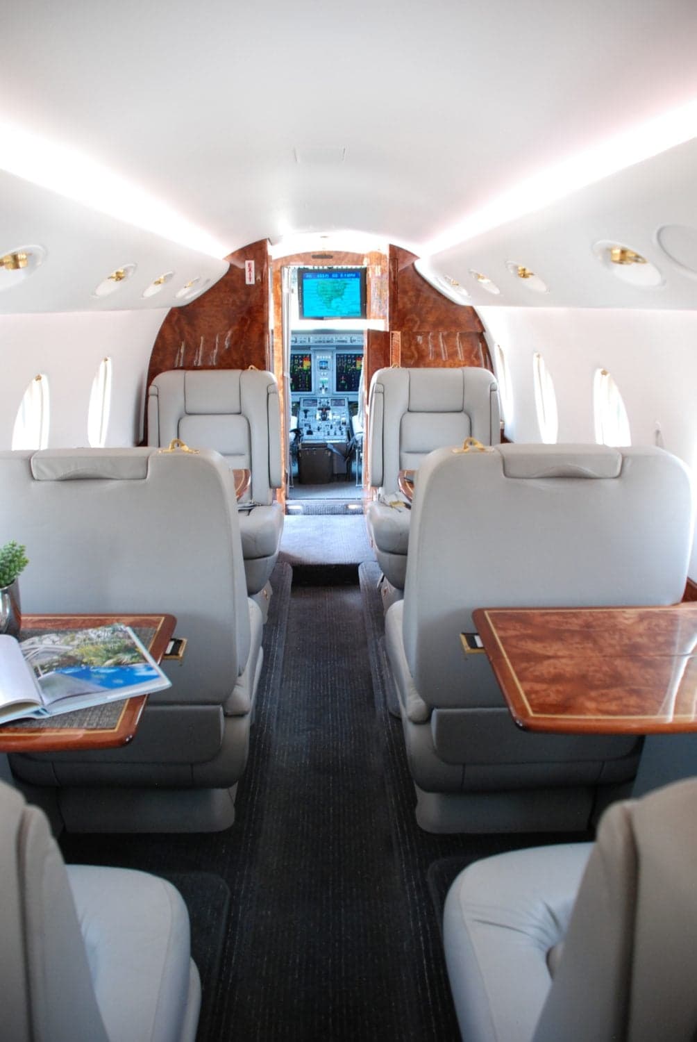 A spacious aircraft cabin with grey leather seats, wood tables, and a view into the cockpit.