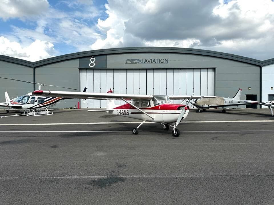 A white and red Cessna 150 G-ASMS parked on the tarmac with a helicopter and another plane, in front of a hangar.
