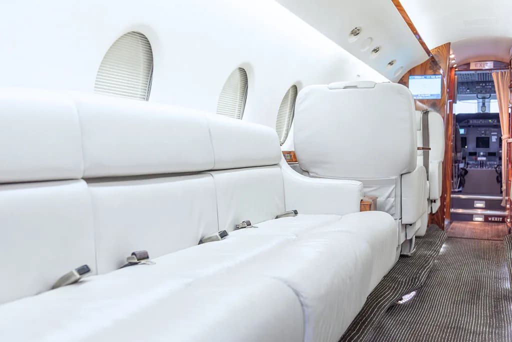 The private jet's cabin interior with white leather seating and a glimpse of the cockpit entrance.