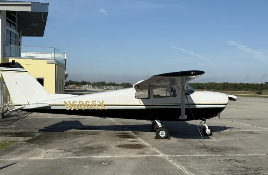 A full side profile view of a white and black Cessna 172 with registration N6865X, parked on an airfield next to a building.