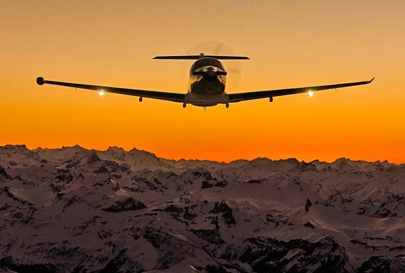 Pilatus PC-12 flying over snow-capped mountains at sunset.