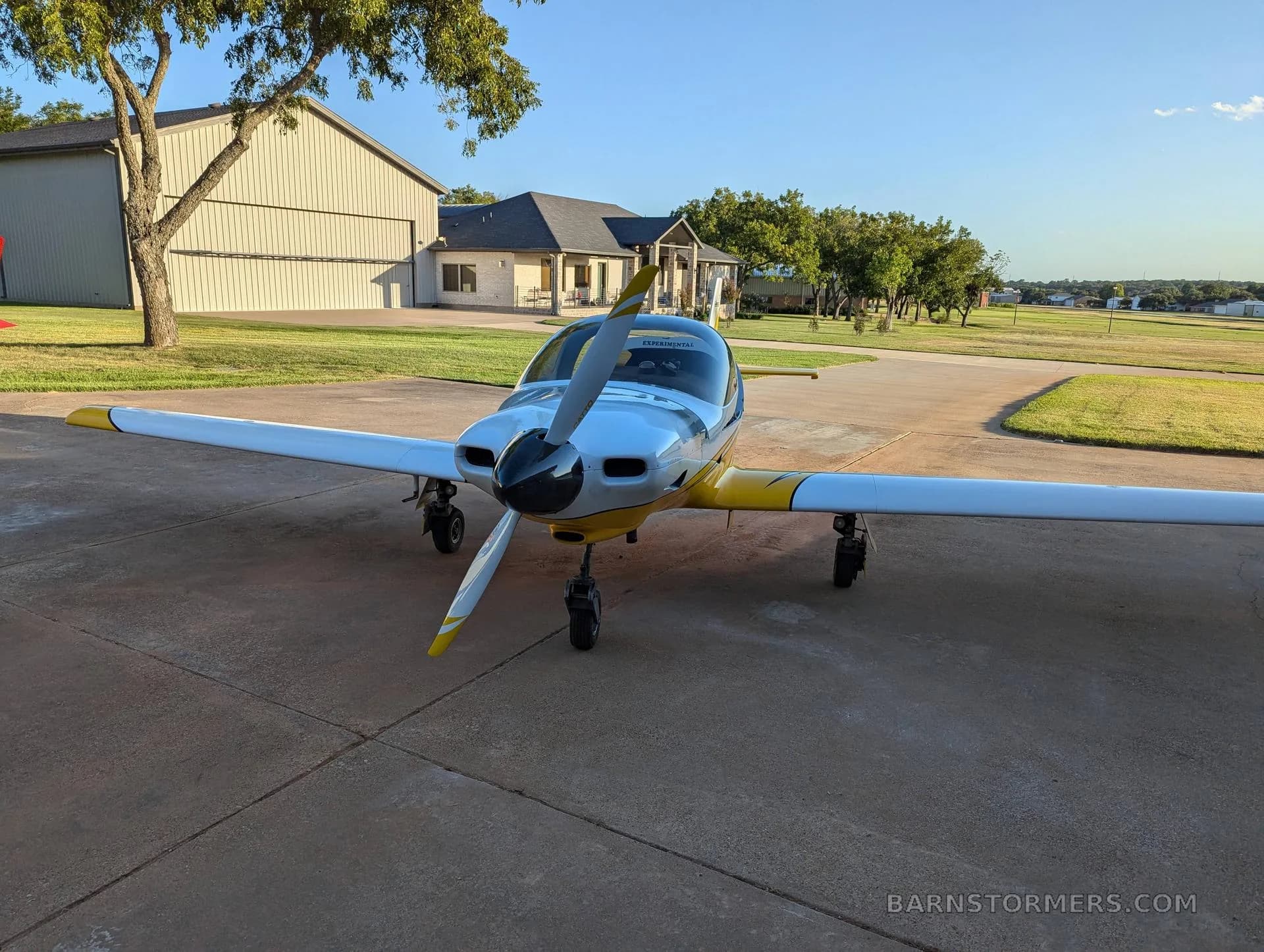 A white and yellow single-engine aircraft with a propeller, viewed from the front, parked on a concrete pad with a building and trees in the background.