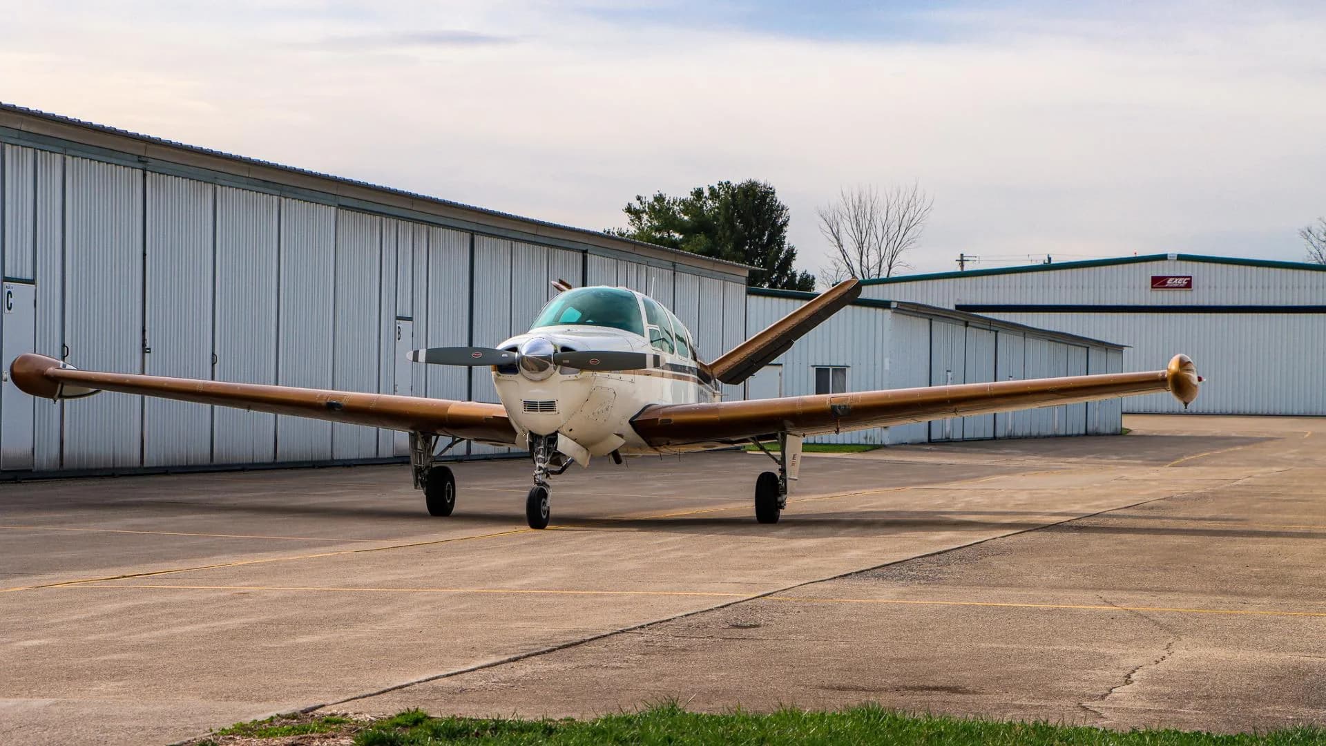 Front three-quarter view of a white and gold Beechcraft Bonanza parked on an airfield with hangars in the background.