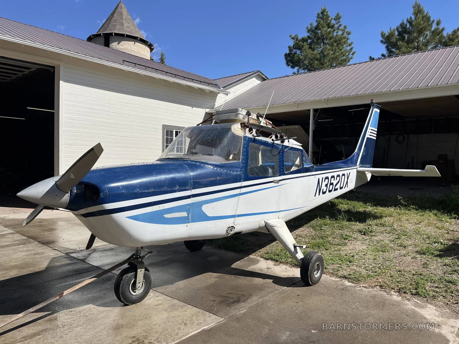 A blue and white single-engine propeller aircraft (N3620X) with a propeller, parked on a concrete pad in front of a hangar.