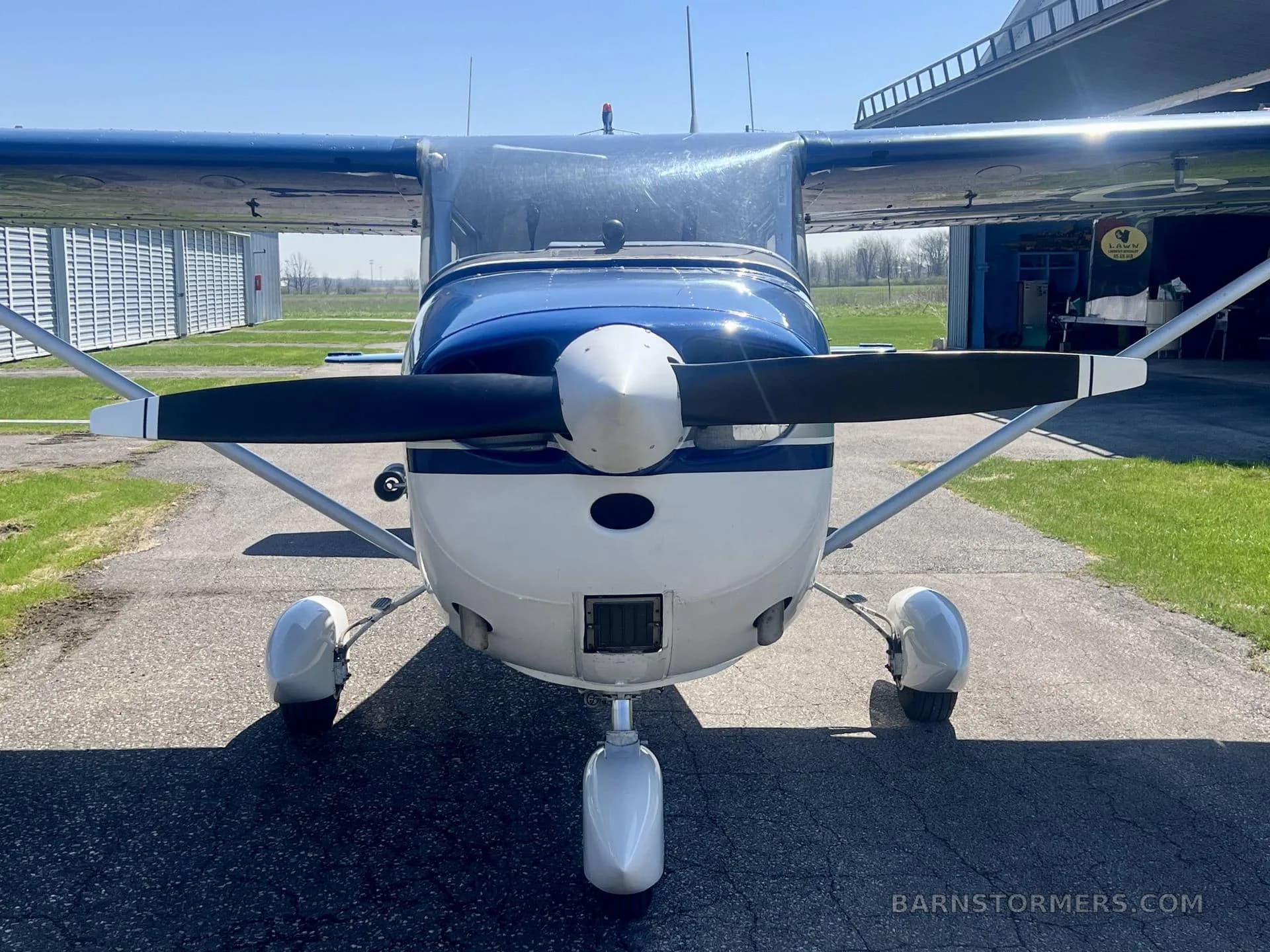 A blue and white 1967 Cessna 172H, registration C-FVKP, parked on an tarmac in front of a hangar, viewed directly from the front.
