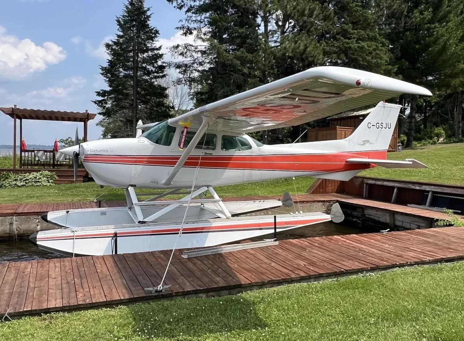 A white and red Cessna Cutlass II floatplane (C-GSJU) is docked at a wooden pier on a sunny day, with trees and a lake in the background.