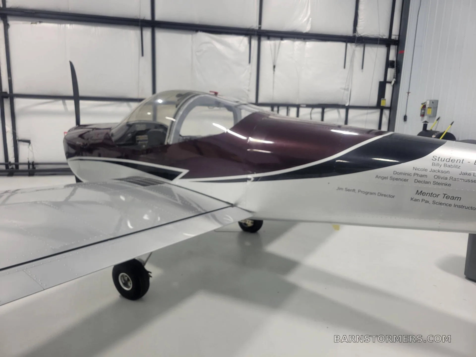 A side view of a burgundy and silver single-engine aircraft, with student and mentor names listed on the fuselage, inside a hangar.