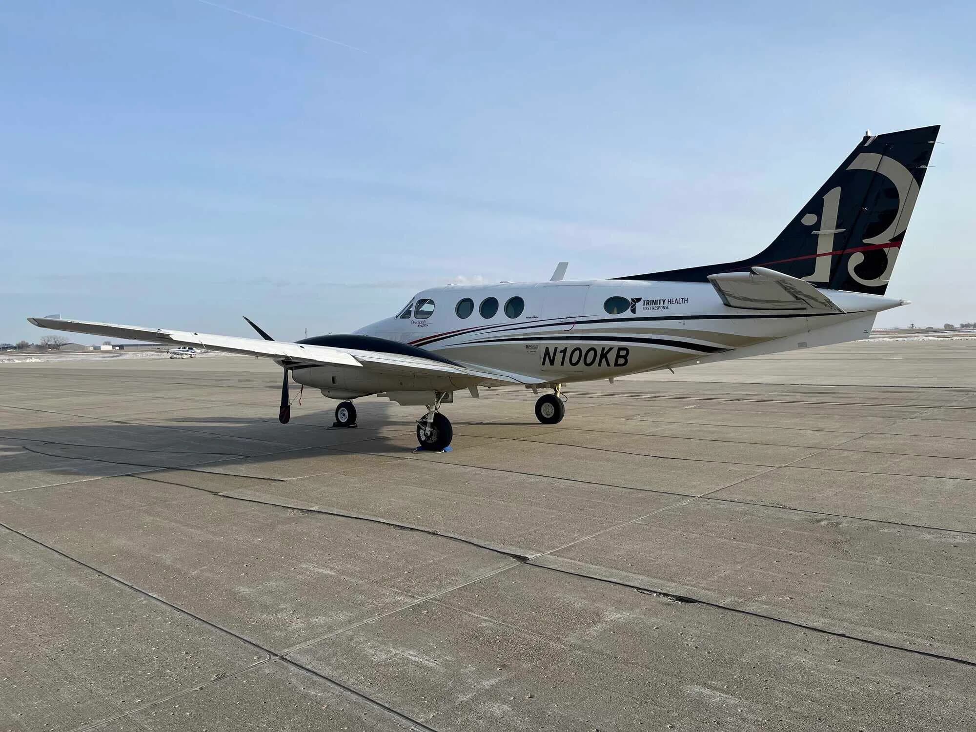 A white and black twin-engine turboprop aircraft with "N100KB" registration and "TRINITY HEALTH" branding, parked on an airfield.