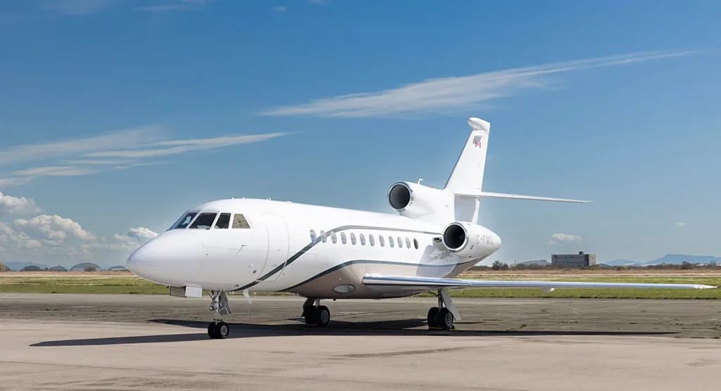 A white Dassault Falcon business jet with a brown stripe, registration C-FMCL, parked on an airfield from a front-side view.