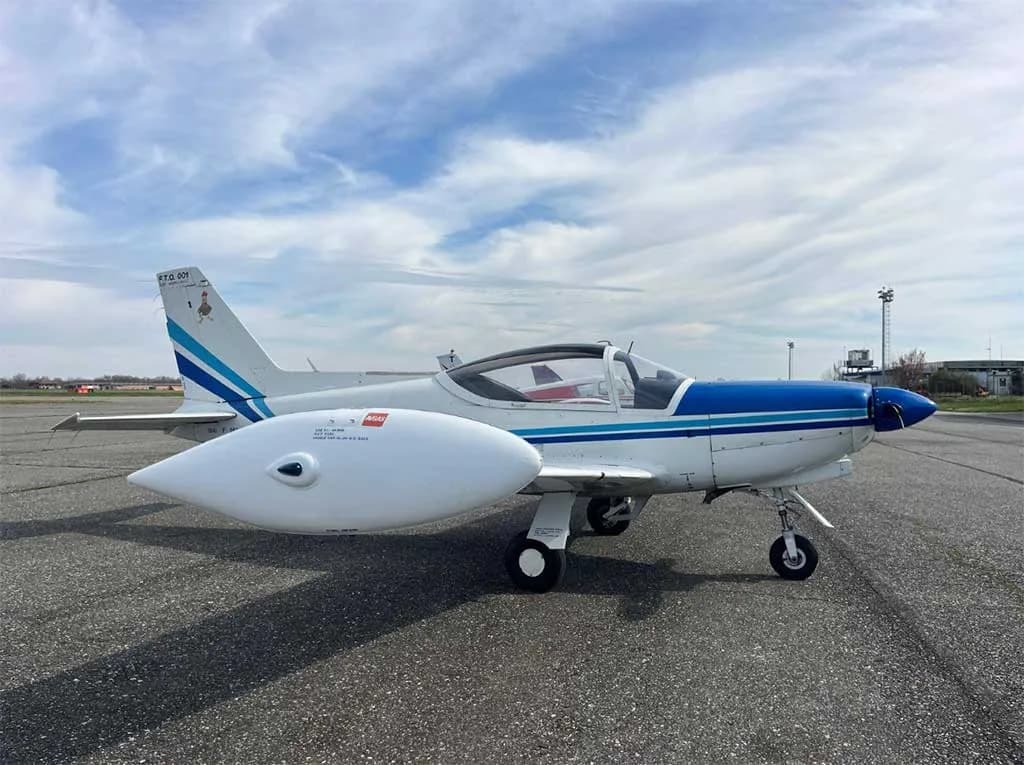 A white and blue single-engine aircraft with large wingtip tanks is parked on an asphalt runway, viewed from the side.