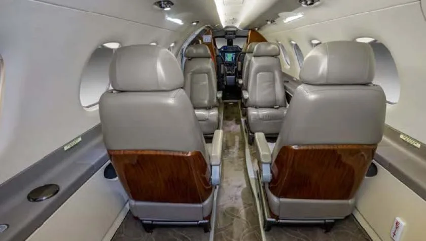 A view down the aisle of a private jet cabin, showing light grey leather seats with wooden backs and a view into the cockpit.