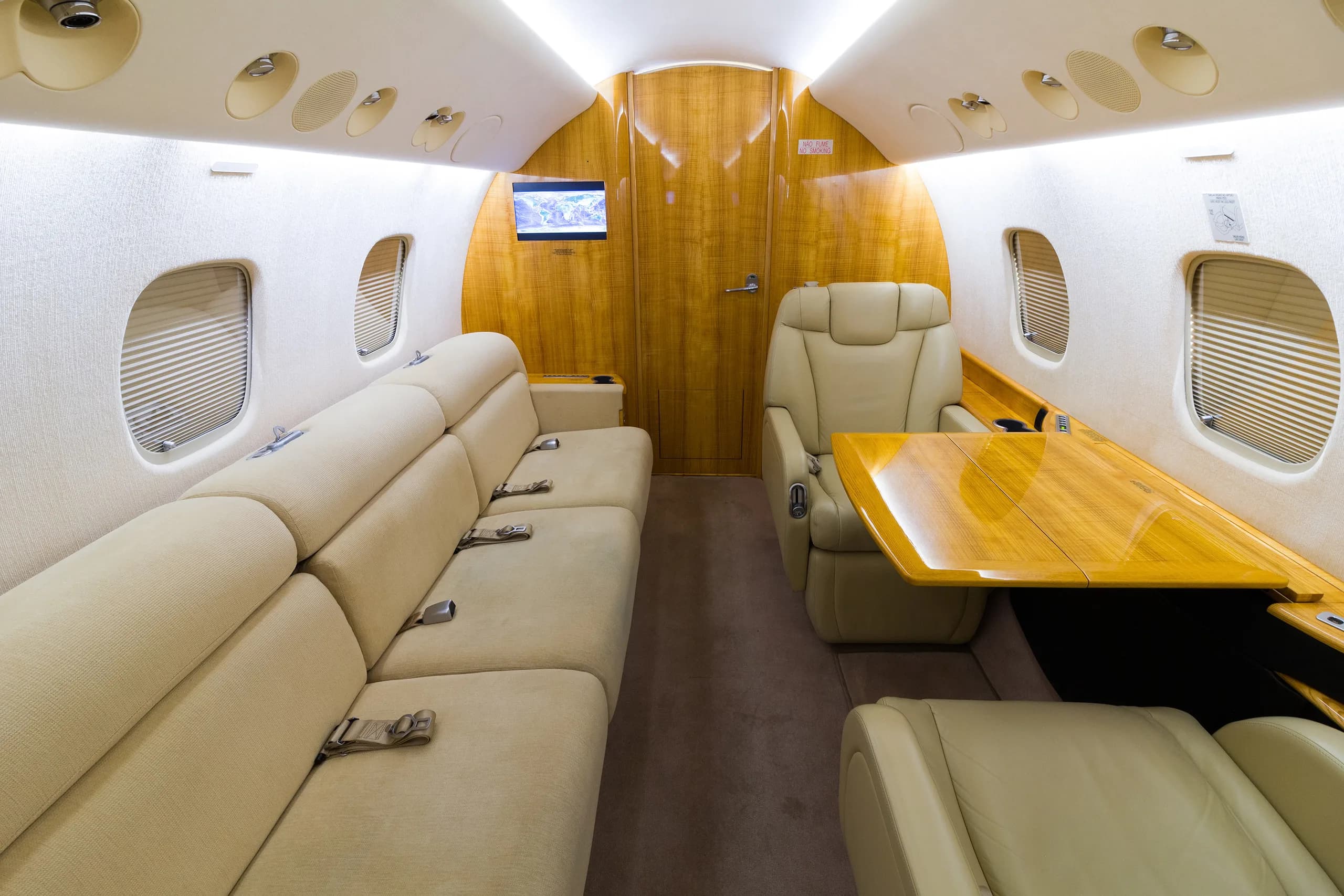 A private jet cabin interior with a long beige sofa, a single beige leather seat, and a polished wooden table.