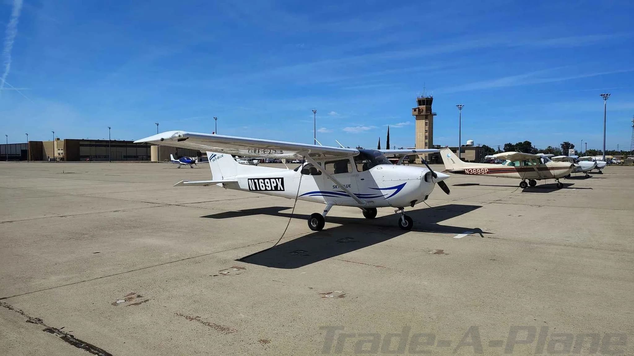 A front-side view of the white Cessna 172, N169PX, parked on an airfield with an ATC tower in the distance.