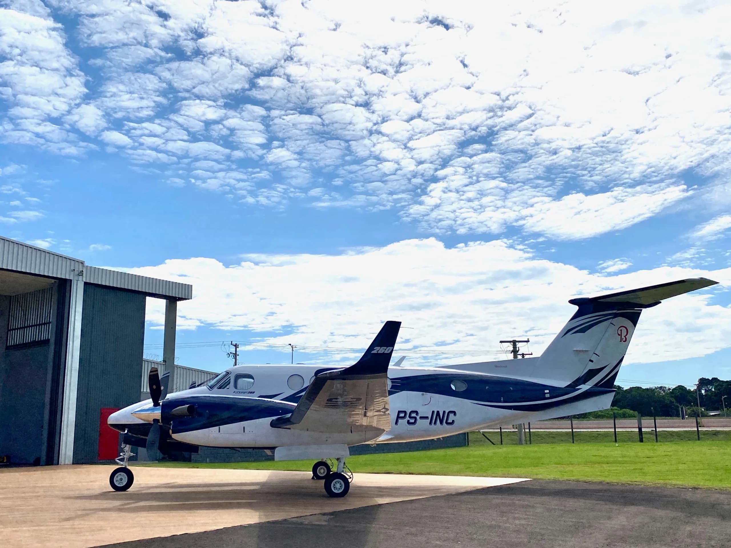 A white and blue twin-turboprop aircraft, registration PS-INC, parked on a tarmac next to a hangar.