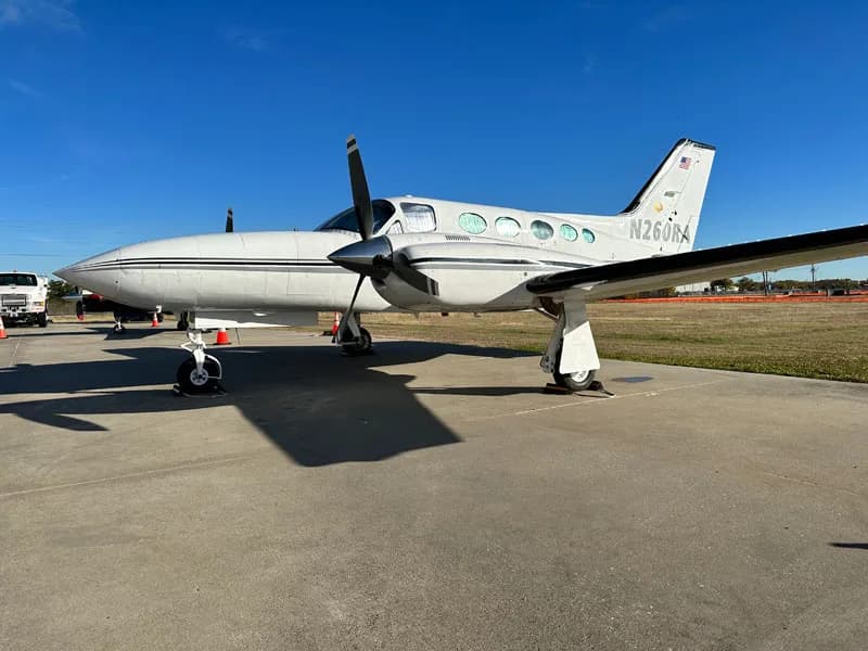 A white twin-engine propeller aircraft with grey stripes, tail number N26JN, parked on an airfield.
