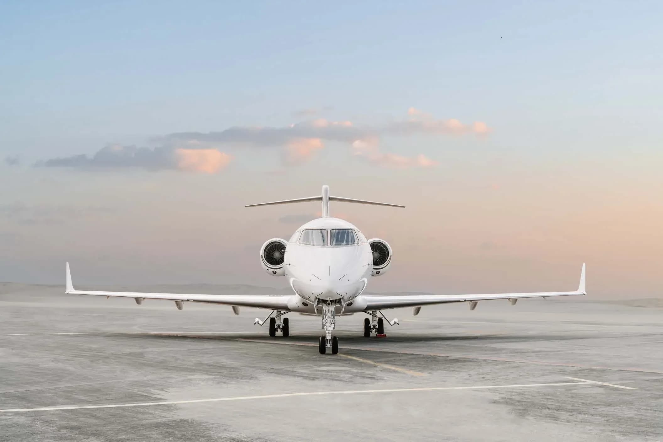 A white business jet with a blue stripe is seen head-on, parked on an airfield under a soft, cloudy sky.