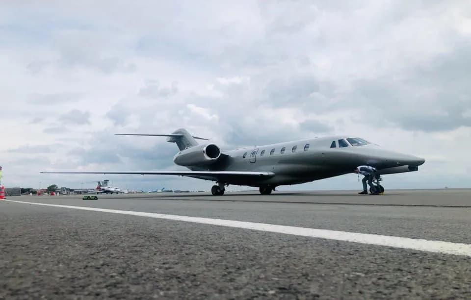 A silver private jet on an airfield with a person near the nose, under a cloudy sky.