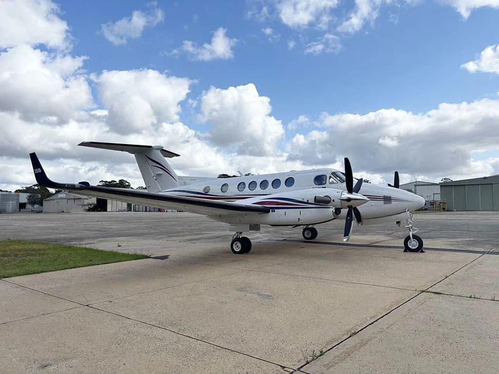 A white twin-turboprop aircraft with red and blue stripes, viewed from the side on a sunny day at an airport.